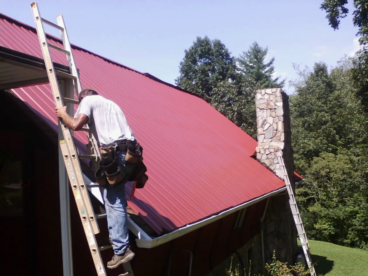 Roofer on a ladder beside a red metal roof and stone chimney — Soffit & Fascia Repair by Roofing Brothers in Mashpee
