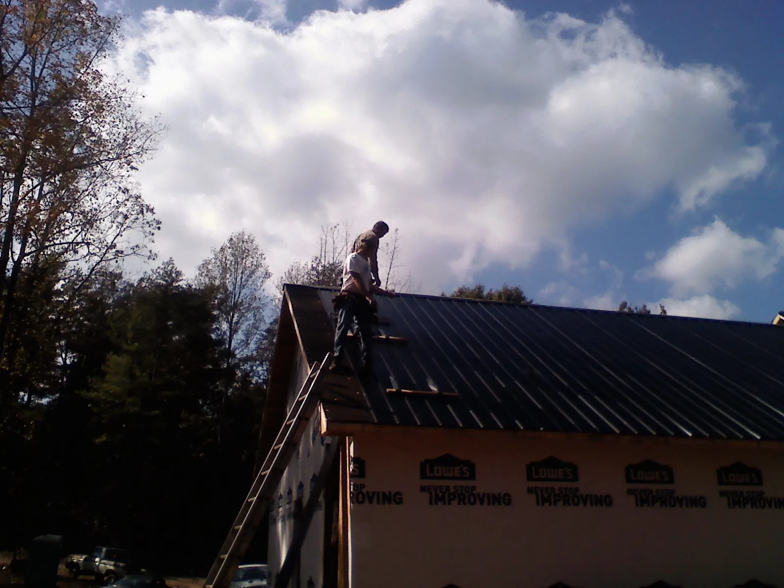 Roofer fastening dark metal panels high against a cloudy sky, from a Roof Inspection job in Mashpee